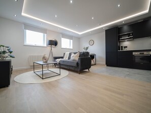 Een moderne woonkamer met een grijze bank, een salontafel en een vloerlamp. Er is een keuken met donkere kasten en een inbouwoven.