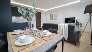 Een moderne woonkamer met een eettafel gedekt voor twee personen, een bank, een televisie en een keuken op de achtergrond.