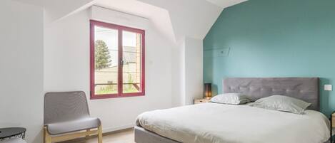 A bedroom with a bed, a chair, a small table, and a window with a view of greenery.