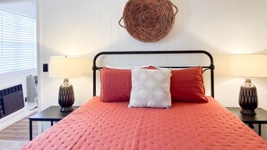 A bed with a red quilt, two nightstands with lamps, a woven wall hanging, and a window with blinds.