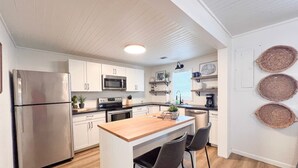 A modern kitchen with white cabinets, stainless steel appliances, a wooden island with bar stools, and decorative woven baskets on the wall.