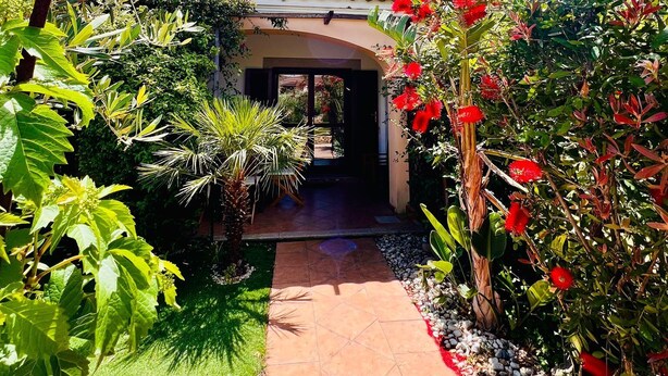 A pathway leading to a building with a tiled entrance, surrounded by lush greenery and red flowers. - Le Querce 5 DX (San Teodoro)