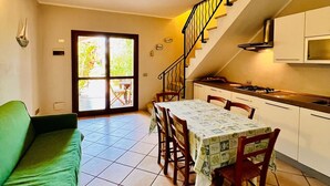 A dining area with a table set for four, a green sofa, a kitchen with white cabinets, and a staircase leading to an upper level. - Le Querce 5 DX (San Teodoro)