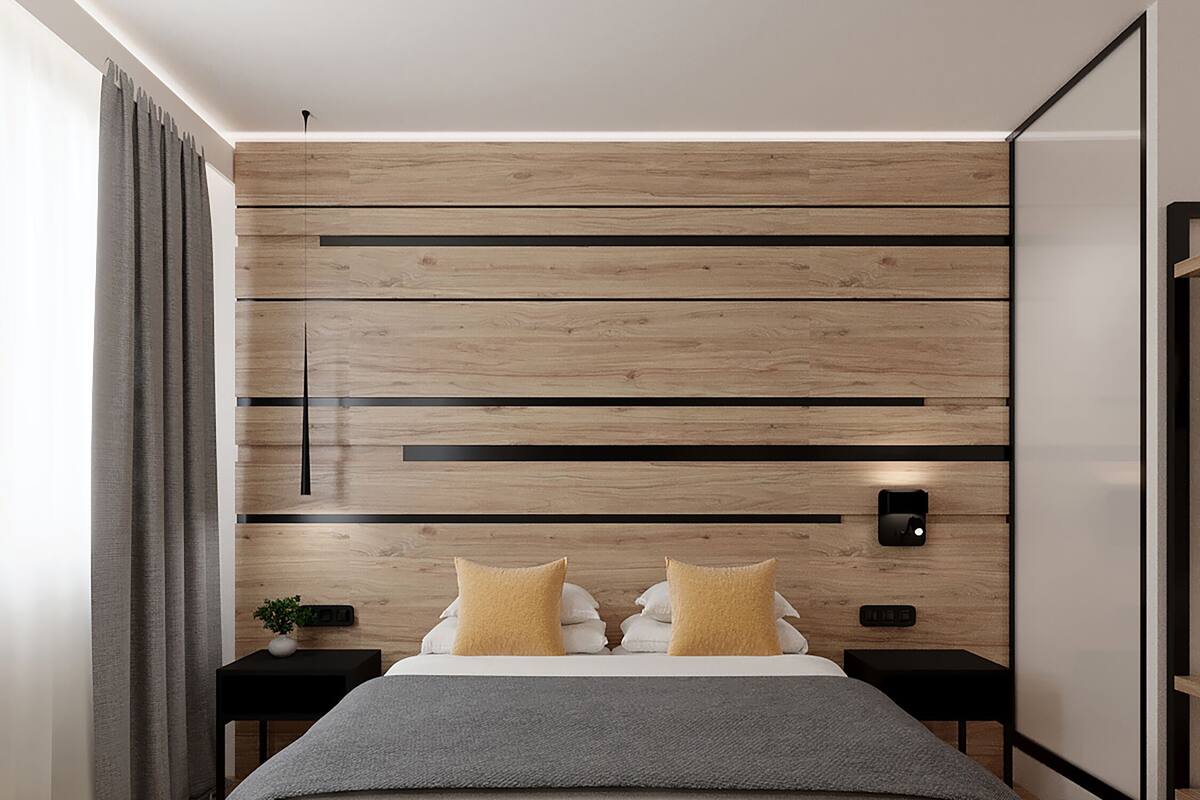A modern bedroom with a wooden headboard, a large bed with white and gray bedding, two bedside tables with lamps, and a view of the outside through a window with curtains.