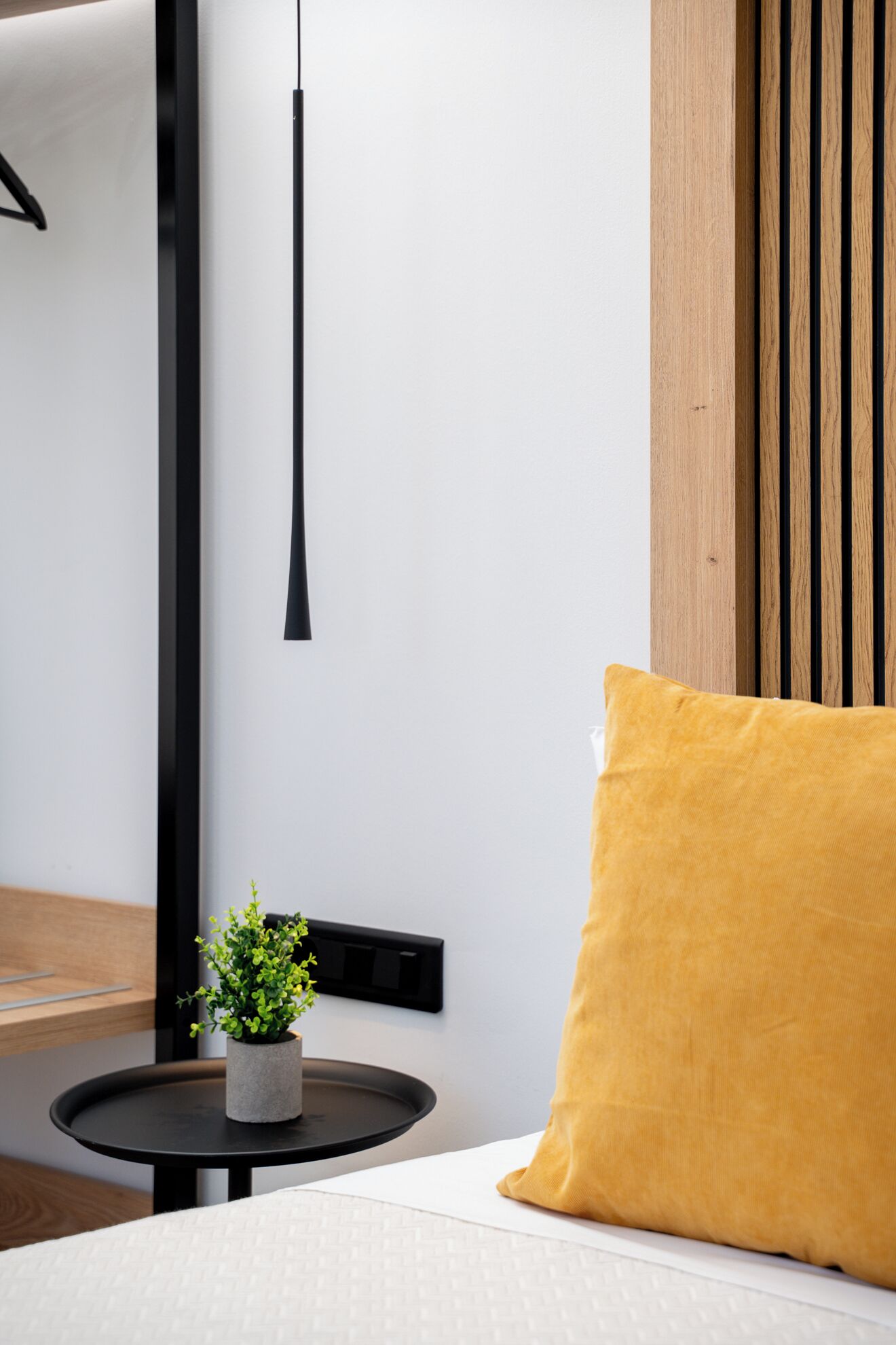 Une chambre d’hôtel bien rangée, avec un lit, une petite table ronde ornée d’une plante et un coussin jaune.