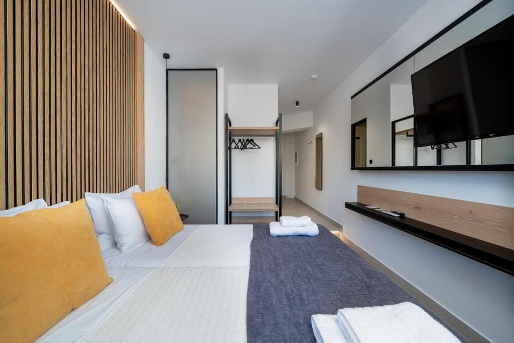 A modern hotel room with a large bed, a flat-screen TV, a wooden accent wall, and a built-in bench.