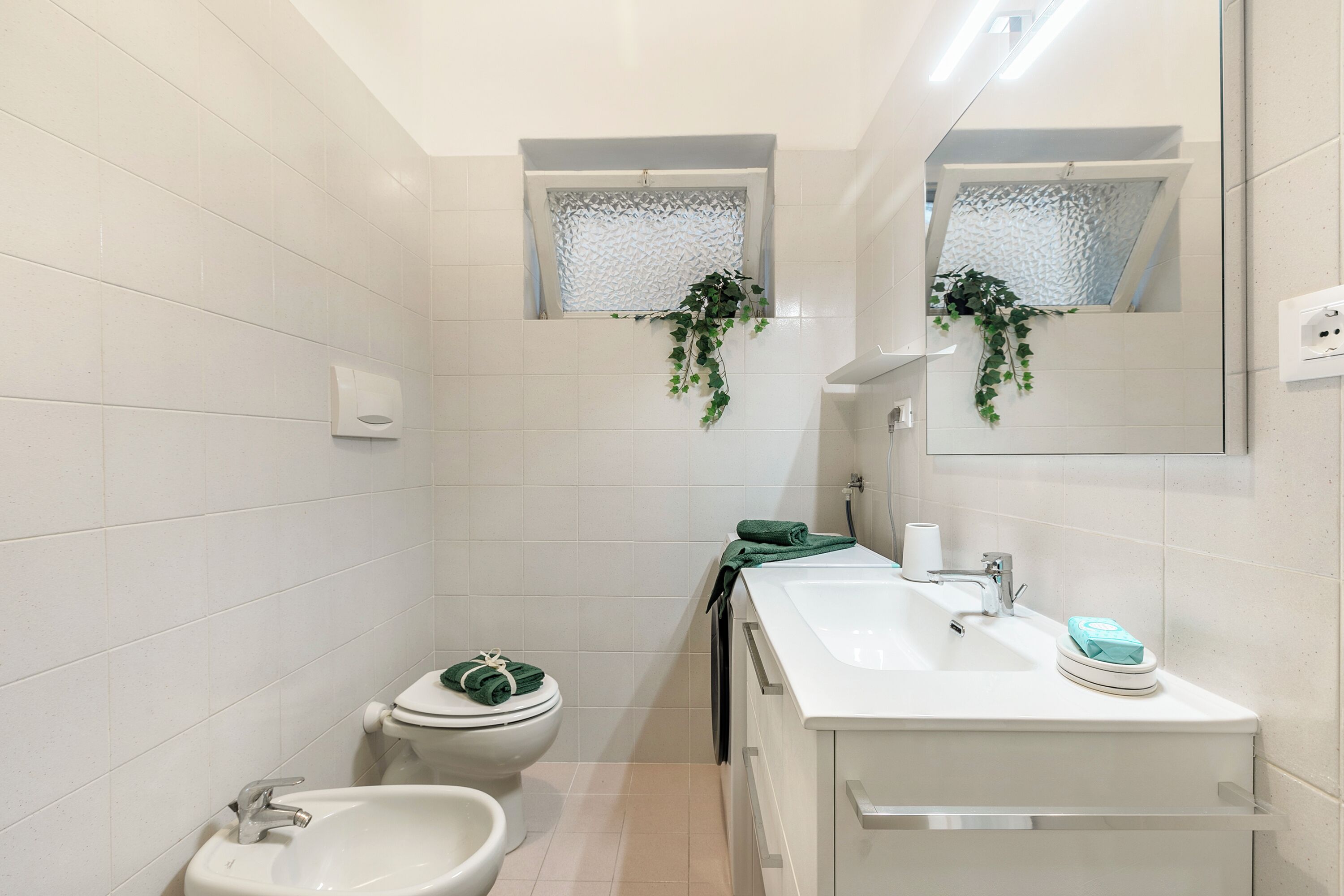 A bathroom with white tiles, a toilet, a bidet, a sink, and a mirror.