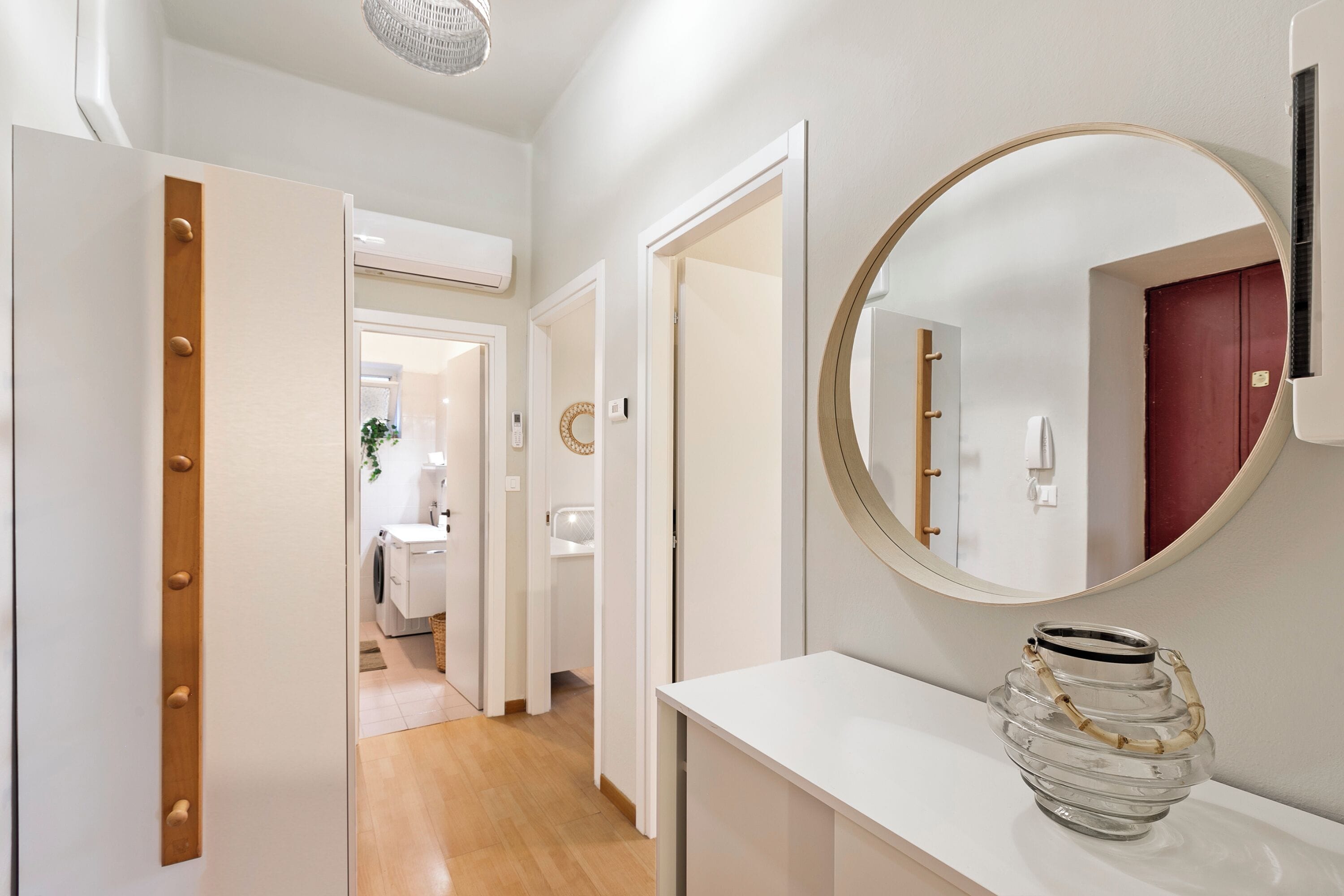 A hallway with a white door, a round mirror, and a glass vase on a white console table.