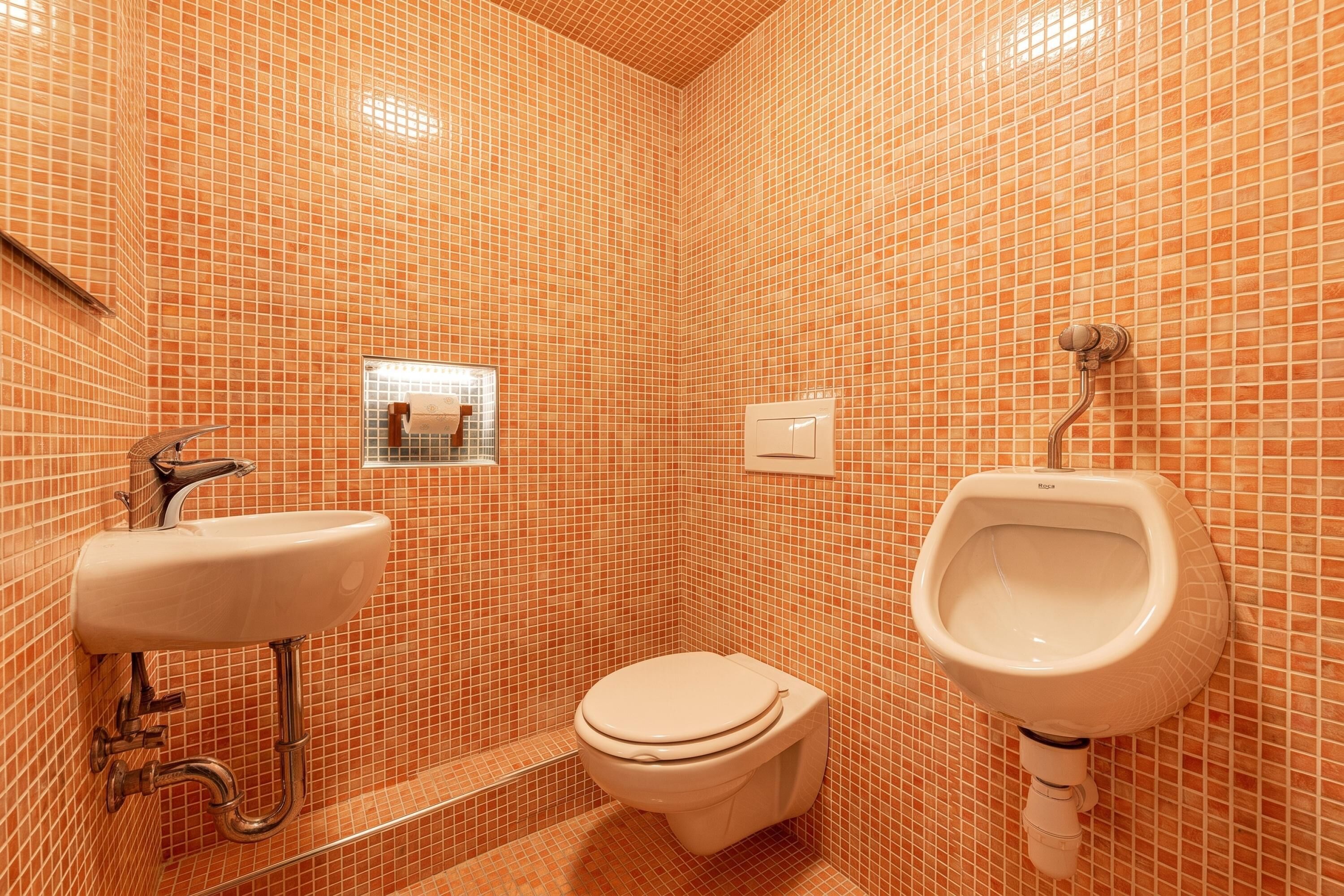Une salle de bain comprenant des toilettes, un lavabo et un urinoir, le tout recouvert d’un carrelage au motif uniforme.