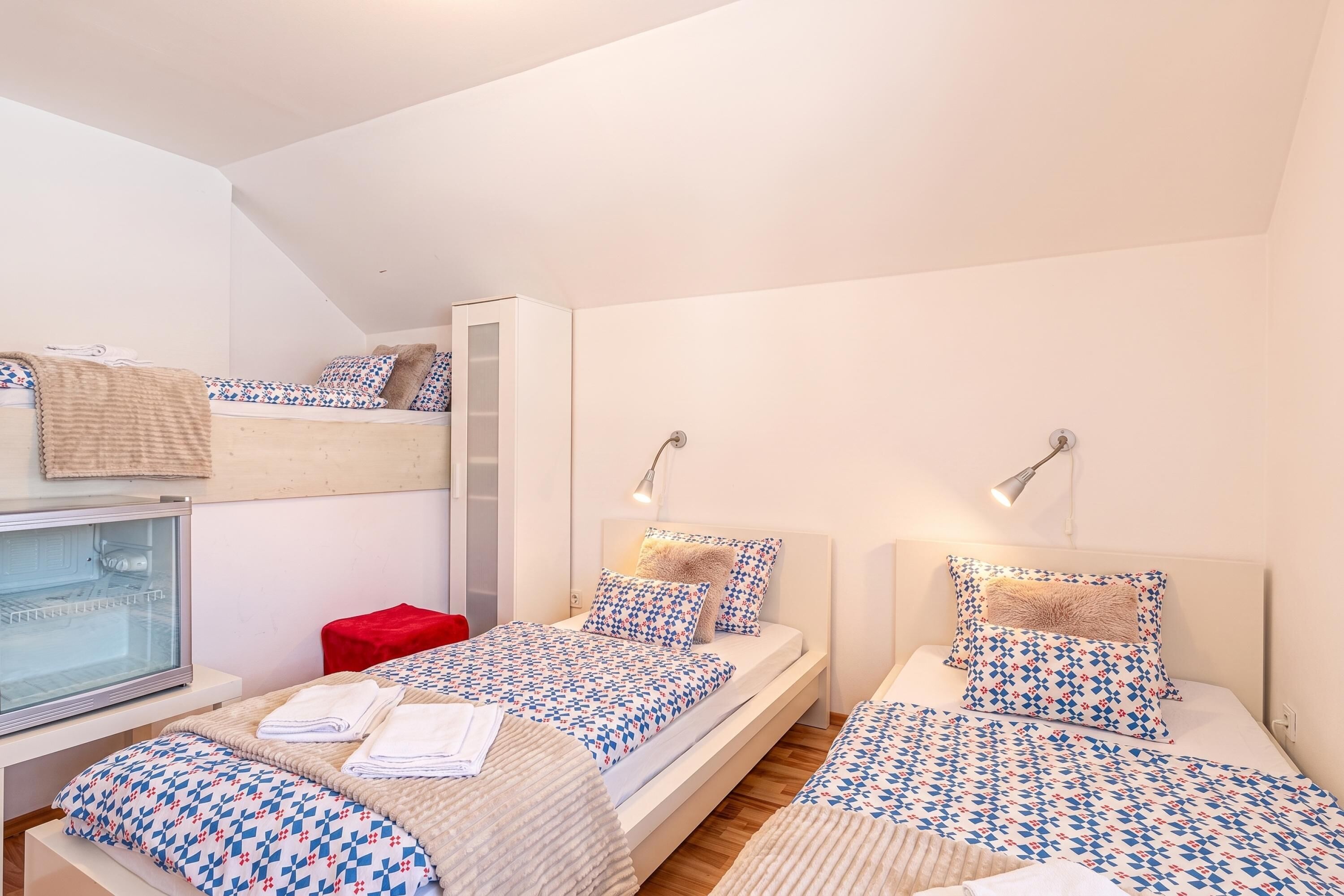 Un dormitorio con dos camas, un refrigerador con puerta de vidrio y un taburete rojo.