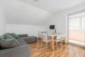 A modern living room with a grey sofa, a dining table with chairs, a flat-screen TV, and a large window. - Apartments Lavrič (Bloke)