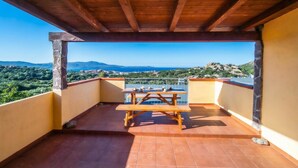 A wooden deck with a table and chairs, overlooking a scenic landscape with mountains and greenery. - Monte Majore 16/b (Olbia)