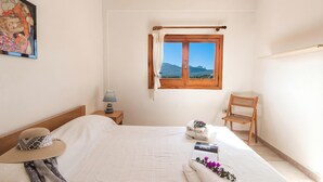 A bedroom with a bed, a window with curtains, a chair, a nightstand, a lamp, and a painting on the wall. - Monte Majore 16/b (Olbia)