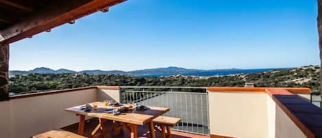 Una mesa de madera y bancos en un balcón con vistas a las montañas y al mar.