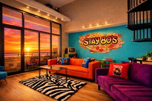 Una sala de estar vibrante con un amplio ventanal que da a un atardecer, un mural de colores y una zona de estar acogedora con un sofá y sillones.