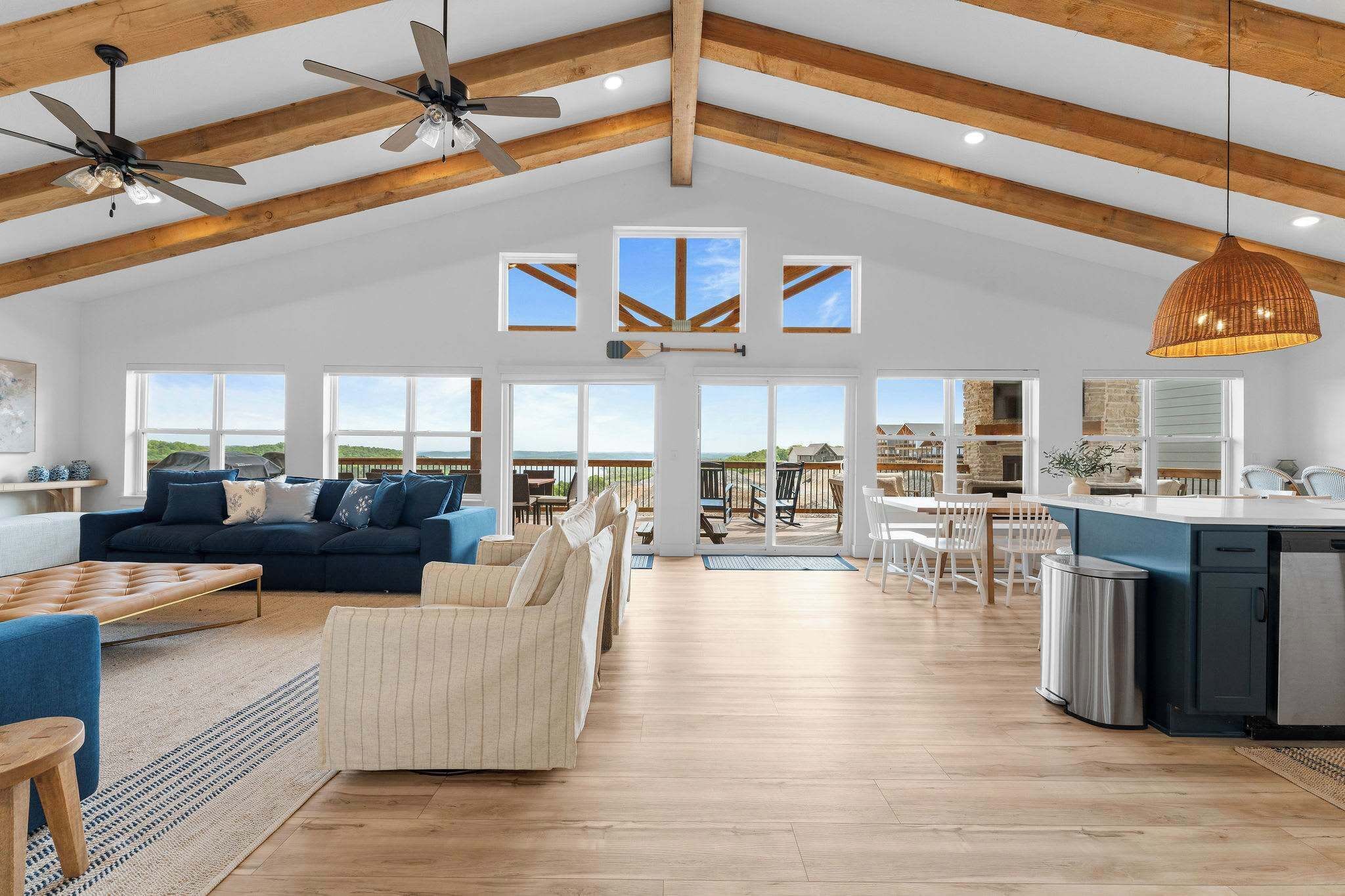 A spacious living area with a high ceiling, wooden beams, ceiling fans, a large sofa, a dining table, and a kitchen area with a sink and refrigerator.