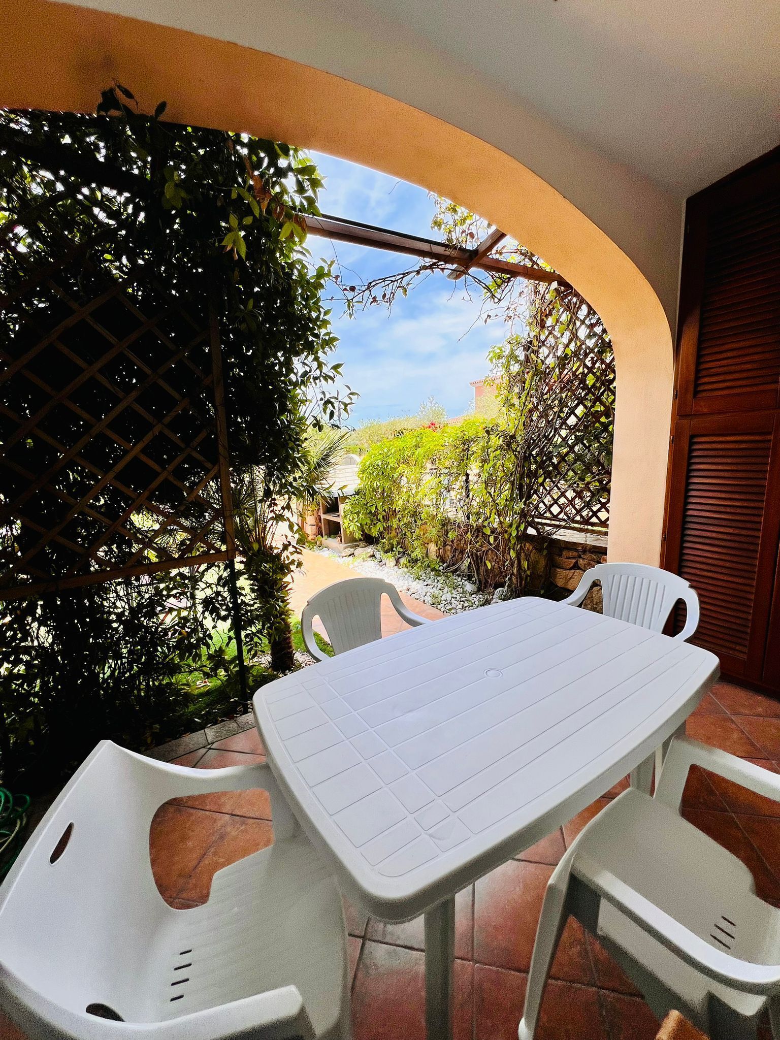 An outdoor patio with a white table and chairs, surrounded by greenery and a clear sky.
