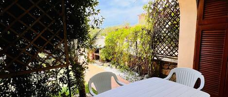 An outdoor patio with a white table and chairs, surrounded by greenery and a clear sky.