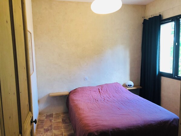 A small, simple room with a bed, a bedside table, a wall socket, and a window with curtains. - Un Coin sur Terre : Lieu Atypique en Pleine Nature (Pougnadoresse)