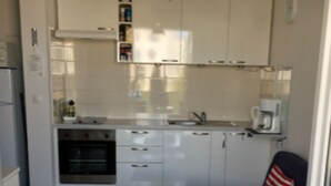 A compact kitchen with white cabinets, a built-in oven, and a coffee maker. There is a small table with a placemat and a dark-colored sofa with a striped pillow. - S&A Apartment (Sutivan)