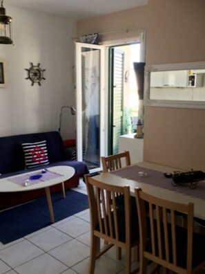 A living room with a sofa, a dining table with chairs, a small kitchen area, and a sliding glass door leading to a balcony. - S&A Apartment (Sutivan)