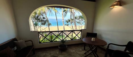 Una habitación con piso de piedra, un ventanal curvo que da a una playa y palmeras, una mesa con sillas y una pequeña planta en maceta.