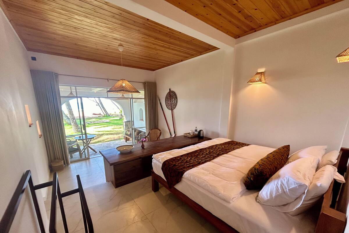 Ein Schlafzimmer mit einem Bett, einem Nachttisch aus Holz, einem Tisch mit einer Lampe und Blick auf einen Garten durch eine Glastür.