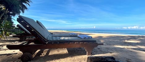 A beachside lounge chair with a towel and a bottle, positioned on a sandy area with a view of the ocean and a palm tree in the background.