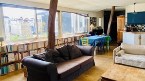 A cozy living room with a sofa, a dining table with chairs, a kitchen area, and a bookshelf filled with books. - Charming flat under the roof (Paris)
