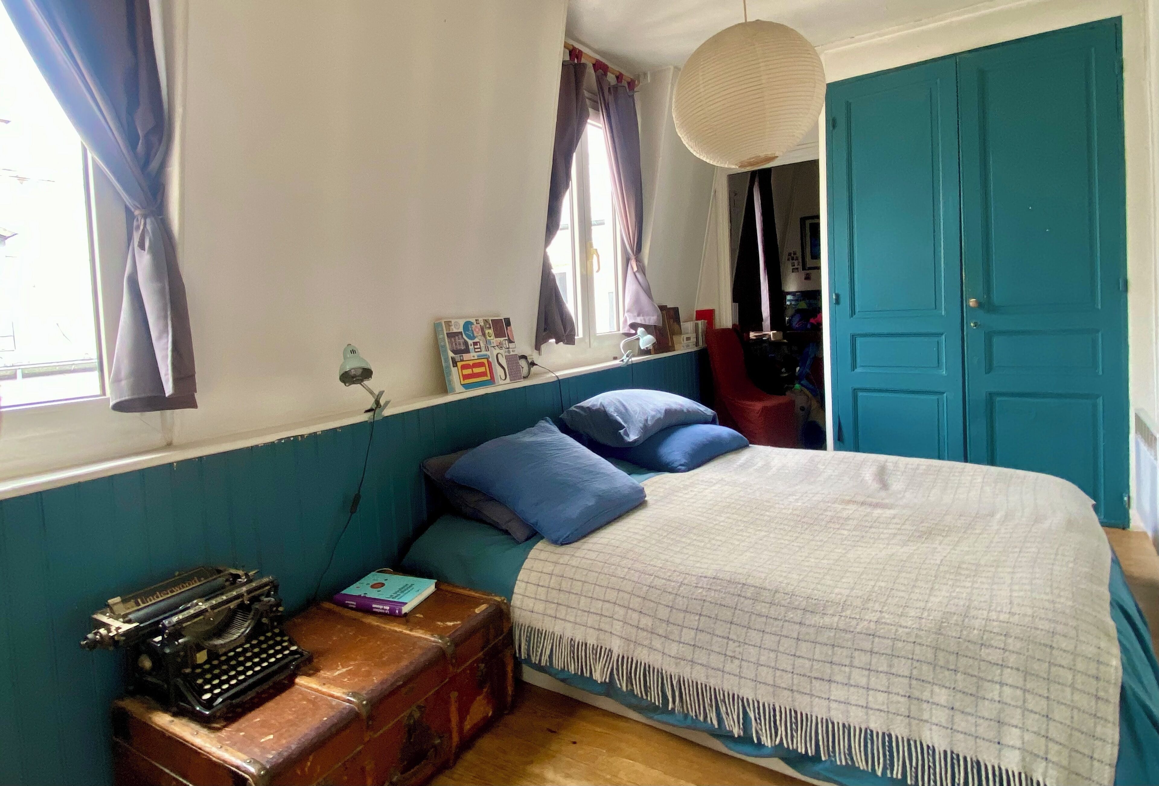 A bedroom with a bed, curtains, a desk with a typewriter, and a suitcase.