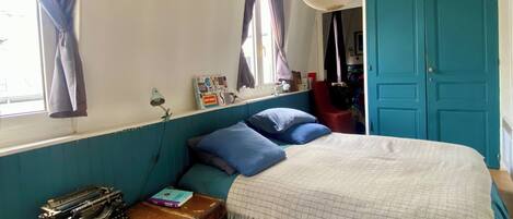 A bedroom with a bed, curtains, a desk with a typewriter, and a suitcase.