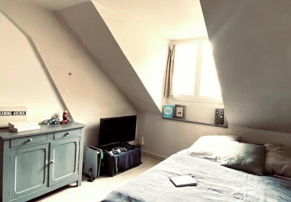A bedroom with a sloped ceiling, a bed, a dresser, a television, and a window.