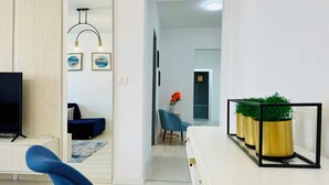 Una sala de estar moderna con televisor, sofá azul y comedor con mesa blanca y plantas en macetas.