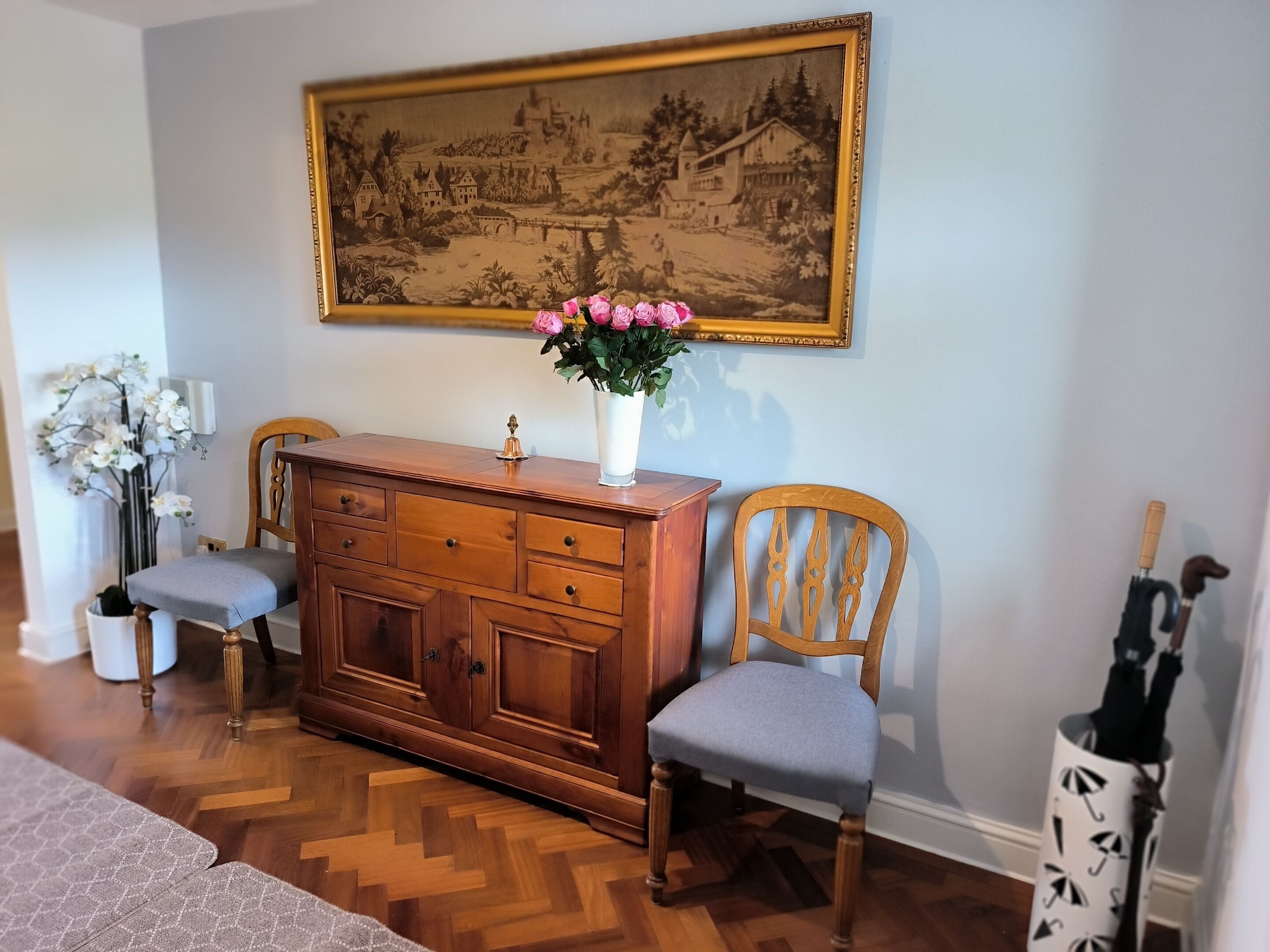 A room with a wooden dresser, two chairs, a painting, and a vase with flowers.