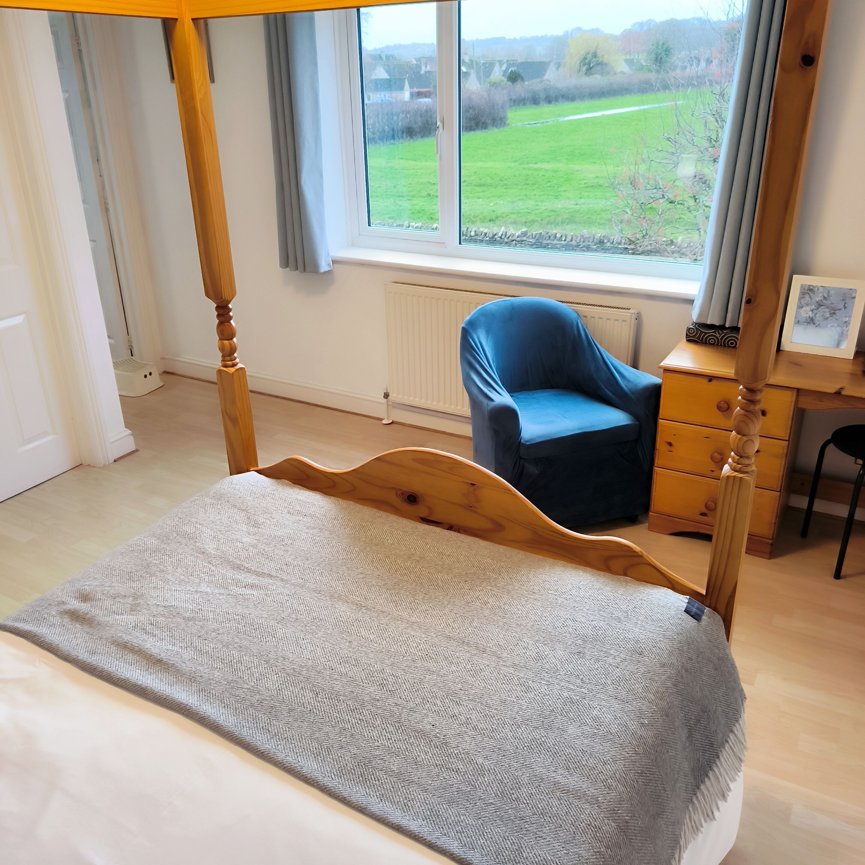 Ein Schlafzimmer mit einem Himmelbett, einem blauen Sessel, einer Kommode und einem Fenster mit Blick auf ein grünes Feld.