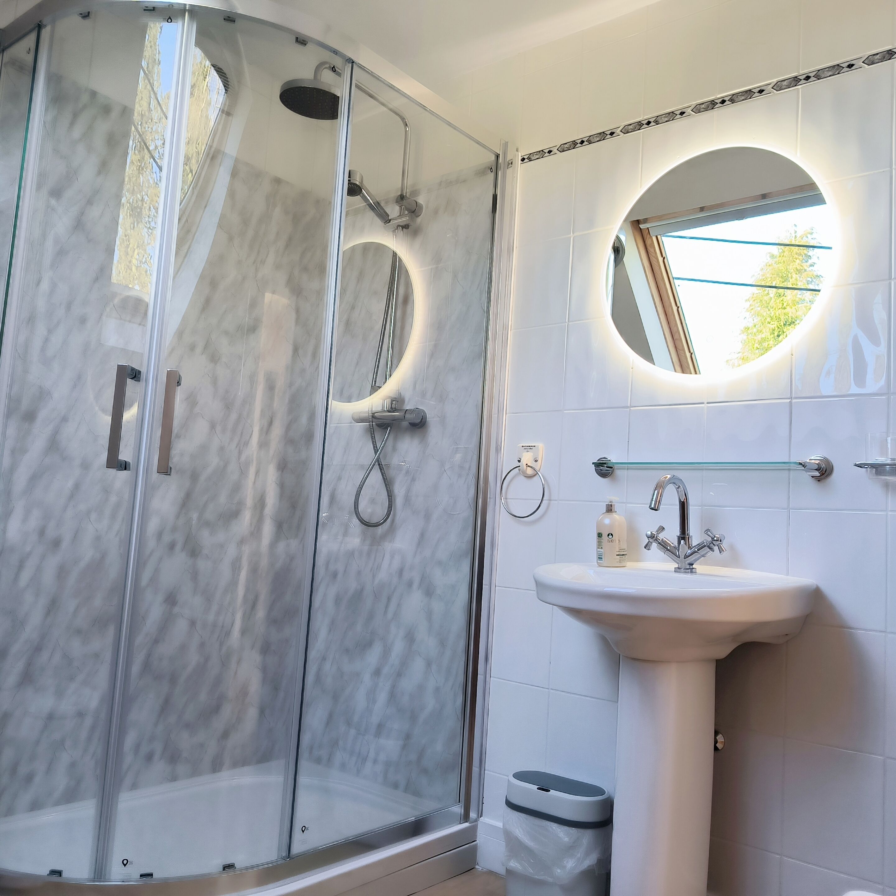 Un baño moderno con cabina de ducha de vidrio, espejo redondo y lavamanos sobre pedestal.