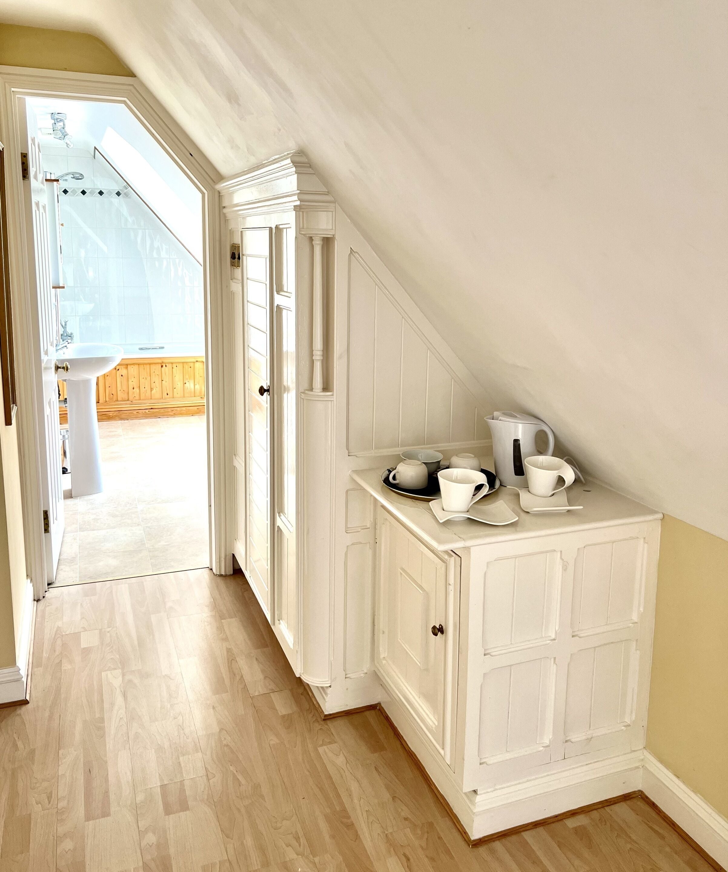A narrow hallway with a white cabinet containing kitchenware, leading to a bathroom with a sink and a wooden floor.