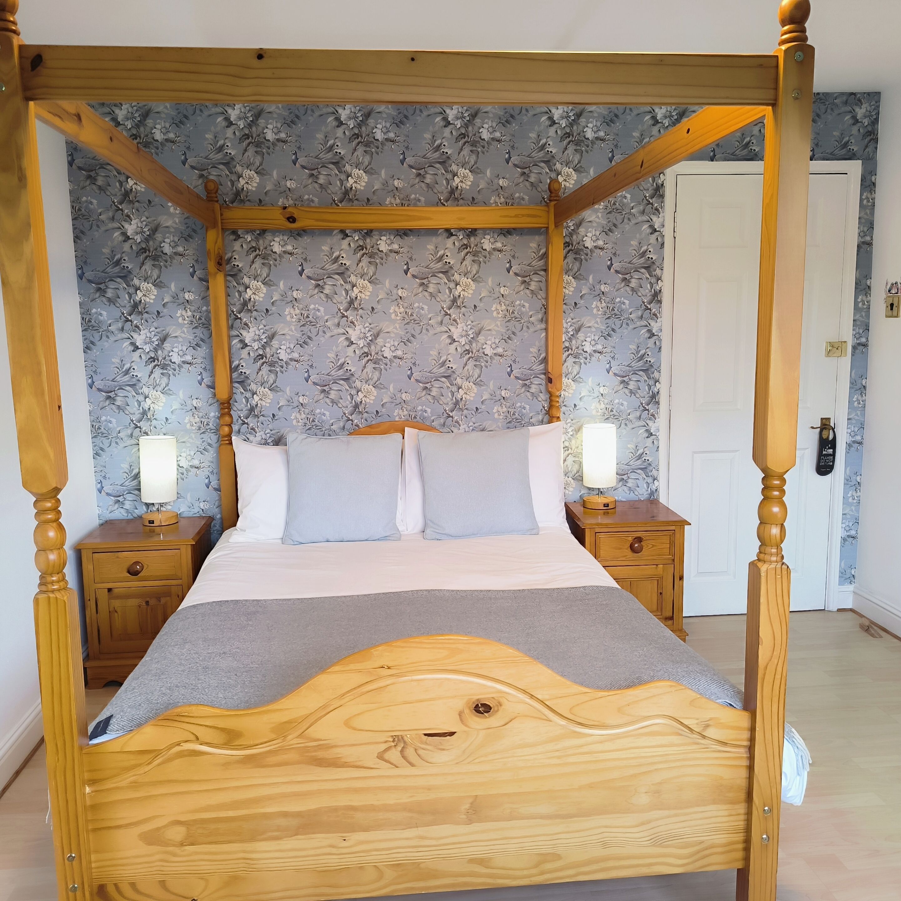A four-poster bed with a wooden frame, two nightstands, and a floral wallpaper background.