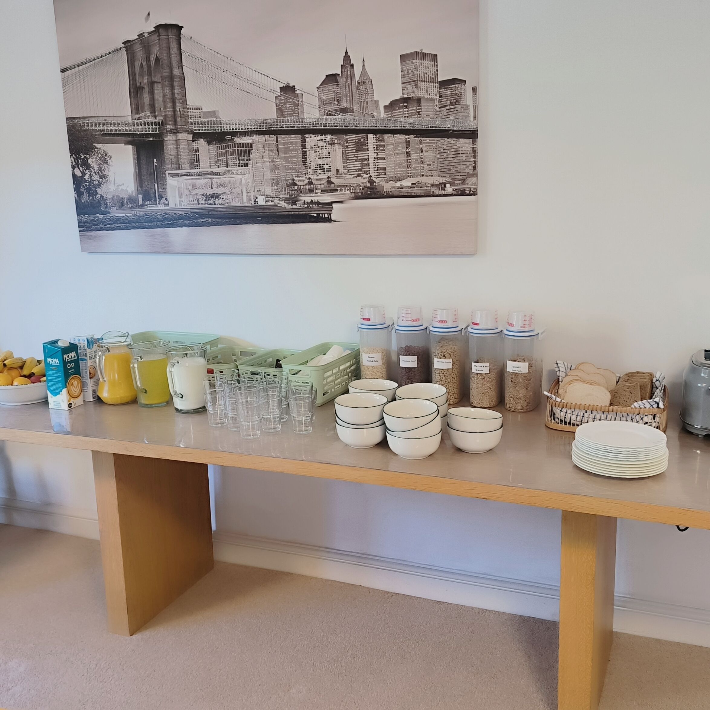 A breakfast buffet table with various food items and containers, a cityscape painting above, and a tiled floor.