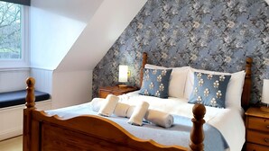 A bedroom with a sloped ceiling, a wooden bed frame, a floral-patterned headboard, and a view of the outdoors through a window. - Meadow Rise (Cheltenham)