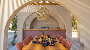 Uma área de refeições com uma mesa de madeira posta para uma refeição, rodeada por arcos e um lustre.