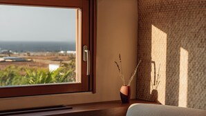 Quarto com escrivaninha de madeira, janela com vista para o oceano e vaso com plantas secas no parapeito.