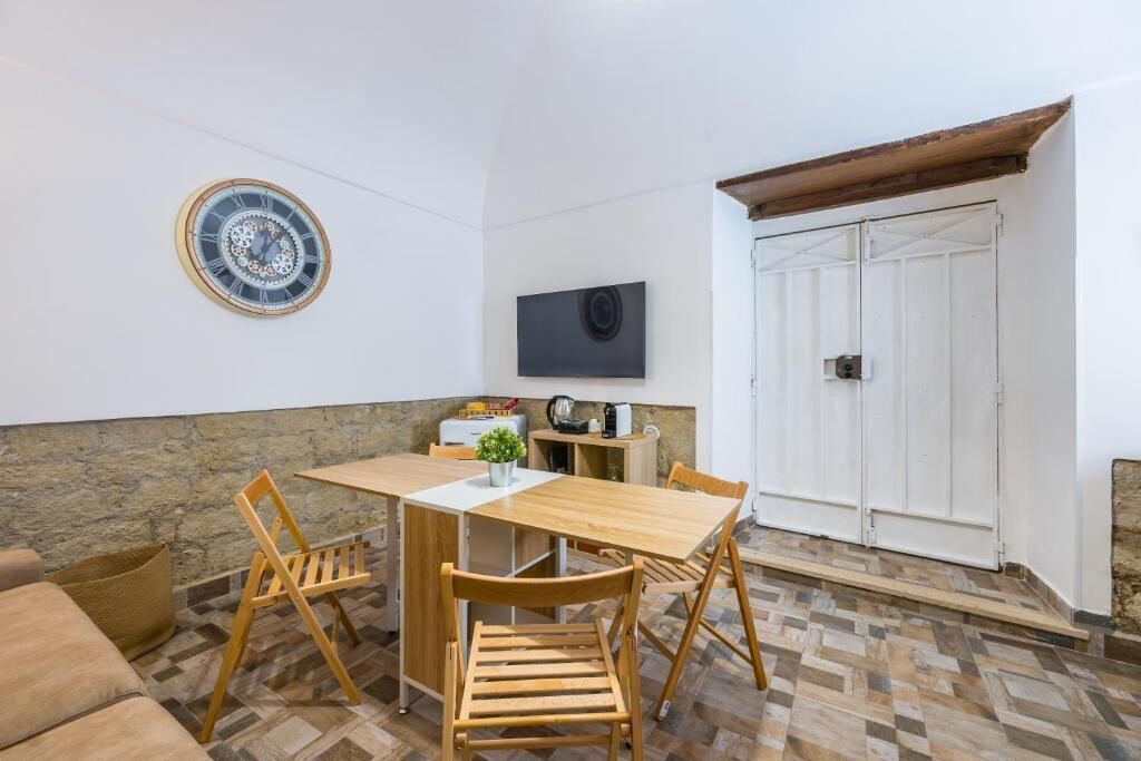 Una habitación con una mesa de madera, dos sillas, una planta pequeña, un reloj en la pared, un televisor y una puerta blanca.