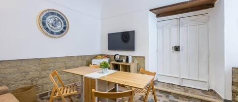 Una habitación con una mesa de madera, dos sillas, una planta pequeña, un reloj en la pared, un televisor y una puerta blanca.