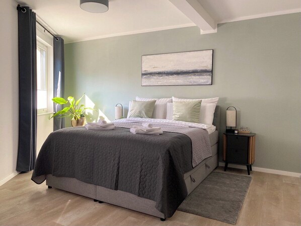 Ein Schlafzimmer mit einem Bett, einem Nachttisch, Vorhängen an einem Fenster und einem Bild an der Wand.