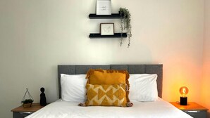 A neatly made bed with a yellow bedspread, white pillows, and a folded white towel on the bed. There are two bedside tables with a small plant on one and a decorative item on the other. Above the bed, there are two black shelves with framed artwork and a hanging plant. - Darlington Suites by SA Stays (Wigan)