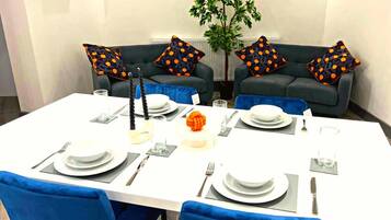 Comedor con mesa blanca dispuesta para cuatro personas, sillas azules y dos sofás azul oscuro con cojines a cuadros anaranjados.