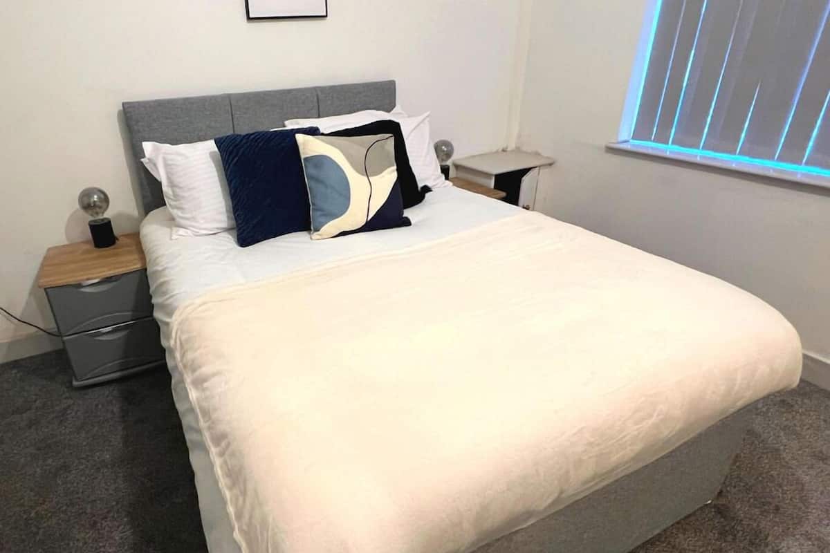A neatly made bed with a gray headboard, white bedding, and a mix of pillows. There are two bedside tables with lamps and a small nightstand. A window with blinds is visible in the background.