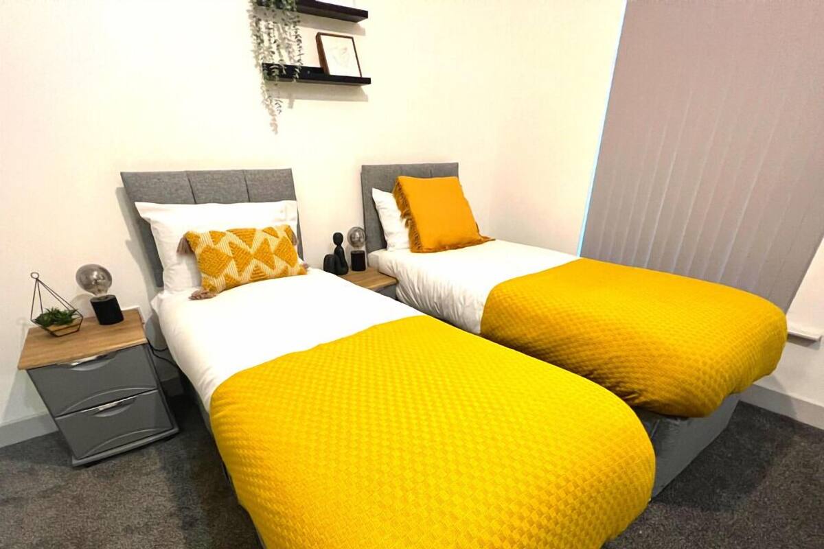 A hotel room with two beds, each with a yellow bedspread and white pillows. There is a small bedside table with a lamp and a plant on the left side.