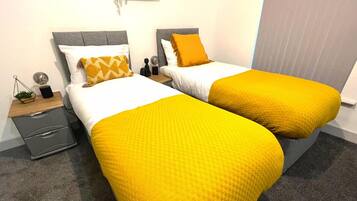A hotel room with two beds, each with a yellow bedspread and white pillows. There is a small bedside table with a lamp and a plant on the left side.